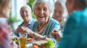 Two visitors sit with an older adult in a bright common room, smiling and chatting respectfully.